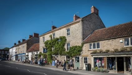 A quaint street scene in a small town with stone buildings, including shops and a pub with ivy climbing its facade. People walk along the sidewalk, and a clear blue sky is overhead.