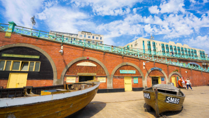 A vibrant promenade scene showing colorful, historic buildings with arched doorways. A sign for the Old Ship Hotel is visible in the background. Two wooden boats are displayed in the foreground, and people are walking along the sunlit path. The sky is bright blue with scattered clouds.