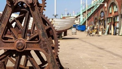 A rusty metal gear mechanism outdoors with a backdrop of a seaside promenade. There are boats, brick arches, and a blue sky in the background. The scene has a maritime, industrial feel.