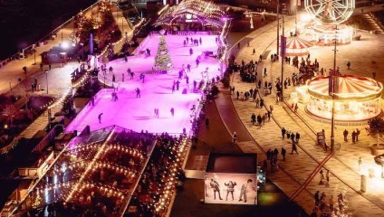 A vibrant nighttime view of an outdoor ice rink illuminated with pink lights, surrounded by festive decorations. Skaters glide on the ice, while nearby crowds enjoy a carousel and Ferris wheel. Warm white lights create a joyful atmosphere.
