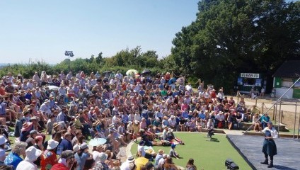 A large crowd sits on tiered grass seating, watching an outdoor theater performance. A person on stage addresses the audience. The setting is sunny with clear blue skies, and trees border the area. Some spectators hold umbrellas for shade. A bar is visible in the background.