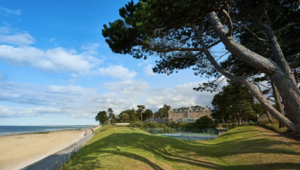 A coastal scene with a sandy beach on the left, a green grassy area with trees in the foreground, and a large building surrounded by more trees in the background. The sky is partly cloudy with patches of blue.