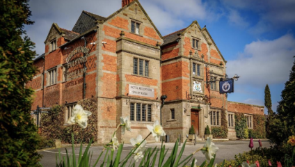 A historic, reddish-brown brick building with a sign for a hotel reception and spa. The building features arched windows and intricate detailing. An emblem sign hangs near the entrance, and daffodils are in the foreground with clear blue skies.