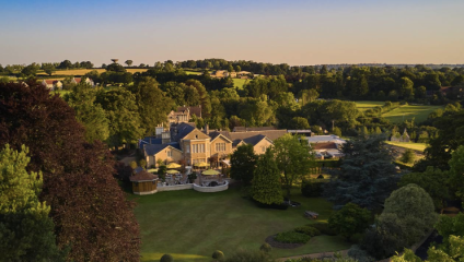 Aerial view of a large estate surrounded by lush greenery and fields. The estate features multiple connected buildings, a well-maintained lawn, and outdoor seating areas. The landscape extends to hills and fields in the background under a clear sky.