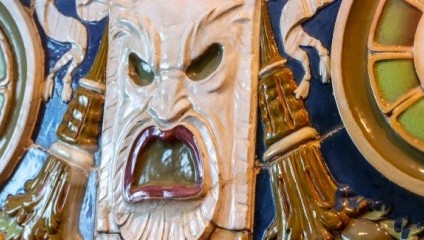 A close-up of a ceramic or stone decorative face with an open mouth and exaggerated features. The eyes are hollow and the expression is intense, surrounded by intricate patterns and colors like blue, green, and yellow.