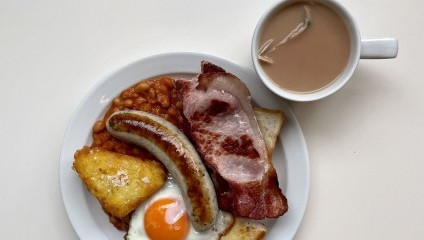 A plate with a traditional English breakfast consisting of a fried egg, beans, sausage, bacon, and a hash brown. Accompanied by a white mug filled with a light brown beverage, likely tea with milk, on a light-colored surface.