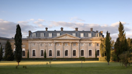 A grand neoclassical-style mansion with tall windows and columns is seen on a clear day. The building features a pediment and symmetrical design. It is surrounded by manicured lawns, tall trees, and bushes, with a blue sky and scattered clouds in the background.
