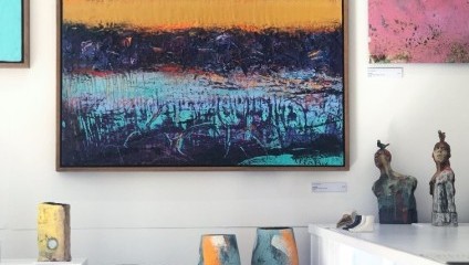 A gallery wall displaying a large abstract painting with bold colors of orange, purple, and turquoise. Below are various ceramic sculptures and vases, including two tall vases with striking blue and orange patterns and two human-like busts with minimalist designs.