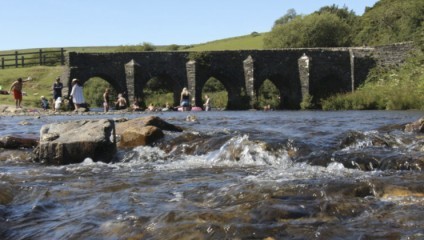 A stone bridge with multiple arches spans over a flowing river. People are gathered at the riverbank, some wading in the shallow water. The surrounding area is lush with green grass and trees, under a clear blue sky.