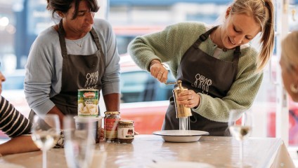 Two women in aprons are engaged in a cooking activity. One woman is using a metal tool to grind or press ingredients onto a plate while the other woman watches attentively. Various jars, cans, and cooking utensils are visible on the table.