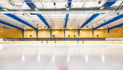A wide shot of the Olympic-sized ice rink at Streatham Ice and Leisure Centre; the pure white ice on the bottom of the screen contrasts with the detail of the blue and yellow rafters high above