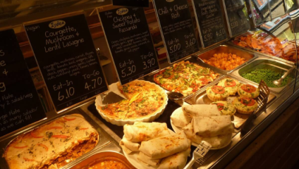 A buffet display featuring various vegetarian and vegan dishes with individual price tags. Items include lasagna, cheese and spinach pasta, chili and cheese roti, a vegetable quiche, burritos, and sides like peas and sweet potatoes.