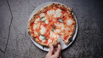 A hand is lifting a slice from a Neapolitan-style pizza on a white plate. The pizza has a charred, bubbly crust and is topped with tomato sauce, melted mozzarella cheese, and fresh basil leaves. The background is a textured, scratched surface.