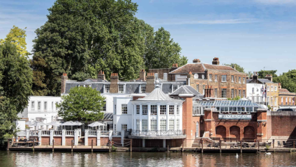 A scenic riverside view featuring historic buildings with brick and white facades, large windows, and rooftop chimneys. Leafy green trees tower behind the structures, and a calm water body reflects the picturesque setting on a sunny day.