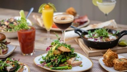 A table set with various brunch dishes and drinks, including a mixed greens salad with avocado and crispy toppings, a Bloody Mary, an espresso martini, a mimosa, a skillet breakfast, and other assorted breakfast items, garnished with fresh herbs and greens.