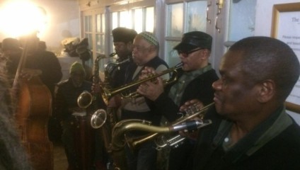 A group of musicians play various instruments in a cozy indoor setting. Four men are playing trumpets, one person is playing a double bass, and another is playing a saxophone. The room is warmly lit, and the musicians are focused on their performance.