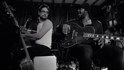A black and white photo of two musicians sitting and playing guitars in a cozy room. The musician on the left, wearing glasses and a long-sleeve shirt, looks at his companion, while the musician on the right, wearing a beanie and a jacket, smiles back.
