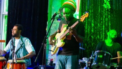 Three musicians perform on stage. The one on the left plays conga drums, the one in the center, wearing a colorful beanie, plays an electric guitar, and the one on the right is on the drum set. The background is dimly lit with green lighting effects.