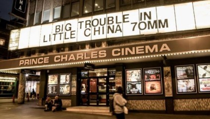 The entrance of Prince Charles Cinema, with a marquee displaying BIG TROUBLE IN LITTLE CHINA 70MM. The facade features posters of various movies, and two people are seated on the ground near the entrance. The area is illuminated with warm lights.