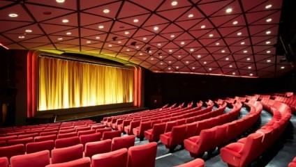 A brightly lit theater interior featuring rows of red upholstered seats arranged in a semicircle. The stage at the front is framed by a red and orange archway and is adorned with closed golden curtains. The ceiling has a geometric design with evenly spaced lights.