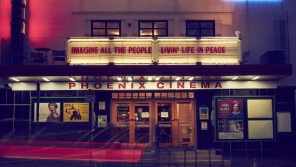 A vintage cinema front at night, illuminated by marquee lights displaying the message: Imagine all the people livin' life in peace. The cinema name, Phoenix Cinema, is visible beneath the marquee. Movie posters are displayed on each side of the entrance.