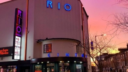 The exterior of Rio Cinema at sunset. The building has a white facade with neon blue letters spelling RIO at the top and CINEMA below. A vertical neon sign reads RIO in purple on the left side. The sky has pink and orange hues, and trees are visible on the street.