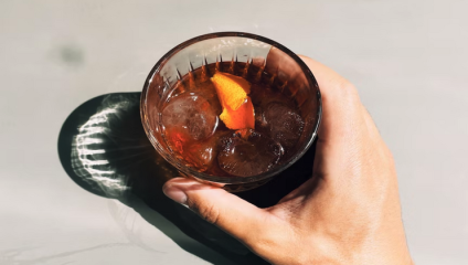 A hand holding a glass filled with a dark liquid, ice cubes, and a twist of orange peel garnish. The glass casts a shadow on the light grey surface below. The scene is brightly lit, emphasizing the clarity of the liquid and the details of the glass.