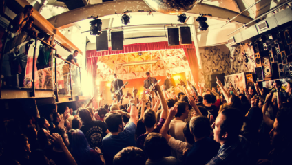 A lively concert with a band performing on stage under bright lights. The energetic crowd, with hands raised, fills the room. The venue features a high ceiling with a disco ball, a mezzanine, and wall decorations. The atmosphere is vibrant and festive.