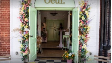 A pastel green storefront with the name Oeuf. The entrance is adorned with colorful floral arrangements. The doors are open, revealing a quaint interior with a person inside. Two benches are positioned outside the shop, and a small sign stands by the door.