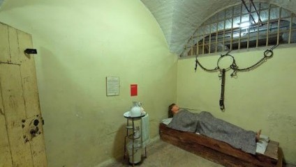 A dimly lit prison cell with arched brick ceiling contains a metal bed with a person covered in a blanket lying on it. Metal shackles hang on the wall to the right. Near the bed is a small table with a white pitcher and bowl. An old wooden door is ajar on the left.