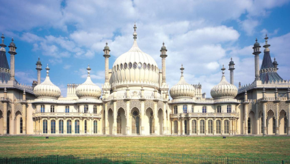 A grand, ornate building with elaborate domes and arches, featuring Islamic and Indian-inspired architecture. The structure is cream-colored with intricate detailing. The foreground showcases a well-manicured lawn under a partly cloudy sky.