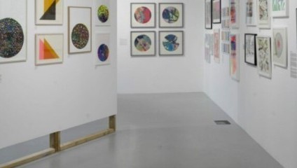 A contemporary art gallery featuring various framed artworks. The walls display colorful abstract paintings, geometric shapes, and intricate designs. The floor is gray, and the room is well-lit, creating a vibrant and inviting atmosphere.