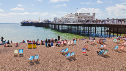 A sunny day at Brighton Beach with people relaxing on the pebble shore. Deck chairs are scattered across the beach, and Brighton Pier stretches into the sea, bustling with visitors. The sky is blue with scattered clouds.