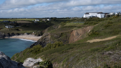 A scenic coastal view featuring a sandy beach surrounded by rugged cliffs and grassy hills. A large white building, possibly a hotel, is situated on top of the cliffs to the right, overlooking the ocean. The sky is partly cloudy, casting a soft light over the landscape.