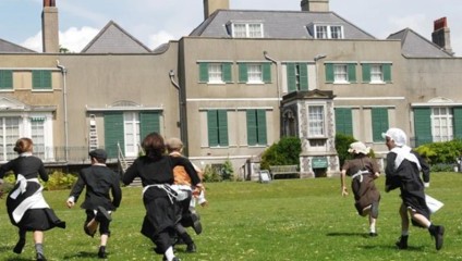 Children in period costumes run across a grassy field towards an old, large, and stately building with green shutters and multiple chimneys. The sunny day adds brightness to the scene.
