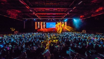 Image of a large indoor conference or event with colorful stage lighting. A sizable audience faces the stage, where a presentation is being shown on a big screen. The venue is dimly lit, with purple and yellow lights illuminating the stage and the crowd.