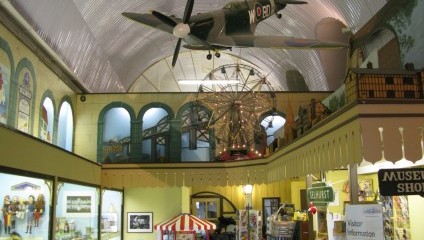 A vintage toy museum interior features a toy biplane hanging from a curved ceiling. Displays of colorful toys, a mini carousel, and a ferris wheel model are visible. The room is filled with vibrant displays and a museum shop on the right side.