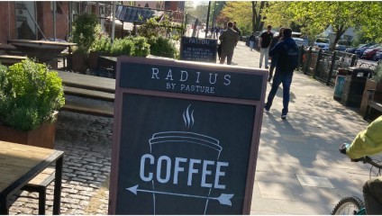 A sidewalk with a chalkboard sign that reads RADIUS BY PASTURE COFFEE featuring an illustration of a steaming coffee cup. The area has outdoor seating with planters, and people are walking and cycling in the background under a canopy of trees.