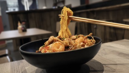 A black bowl filled with stir-fried noodles, shrimp, and vegetables sits on a wooden table. A pair of chopsticks is lifting some noodles from the bowl. The background shows a blurred interior of a restaurant with tables and chairs.