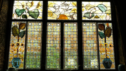 A large stained glass window divided into four vertical panels. The top sections feature colorful floral and butterfly designs. The bottom sections have intricate grid patterns with star-shaped decorations and display potted plants at the base.