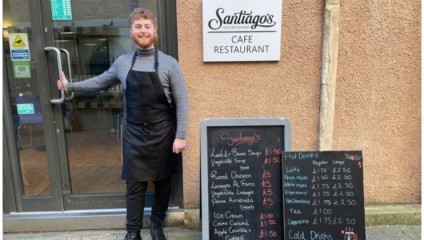 A young man with red hair and a beard smiles while standing outside Santiago's Cafe Restaurant. He is wearing a grey turtleneck, black apron, and black pants, and is holding the door open. A chalkboard menu next to him lists soups, meals, ice creams, and drinks.