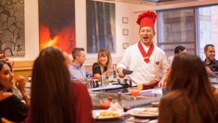 Man in red chefs hat and whites cooking in front of diners