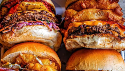 A close-up of four large, stacked burgers. Each burger is overflowing with toppings, including cheese, vegetables, and other ingredients. The buns are golden and slightly toasted. The variety of ingredients creates a colorful and appetizing display.