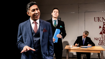 Three men are in a formal setting. The man in the foreground is speaking and wearing a blue suit. Behind him, the second man in a suit holds blue folders. The third man sits at a desk, writing. A logo and the text UK National News Group are visible on the wall.