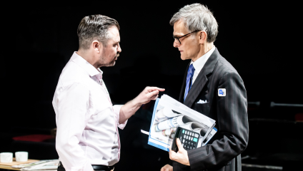 Two men engaged in a serious conversation. The man on the left is gesturing with his hand, wearing a light-colored shirt. The man on the right is holding a folder, rolled-up documents, and a calculator, dressed in a dark suit with a blue tie and nametag.