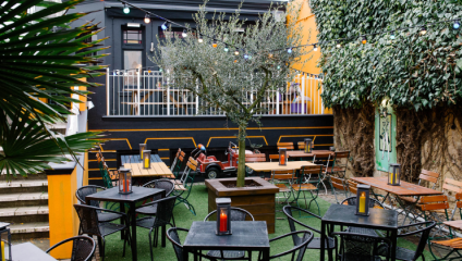 Outdoor patio with various tables, chairs, and string lights hanging above. There is a large tree in the center and greenery climbing up the walls. Some tables have candles or lanterns on them. In the background, a small red toy car is parked near a railing.