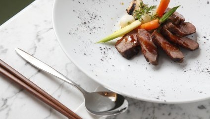 A plated dish featuring several pieces of glazed meat arranged in a semi-circle with assorted vegetables, such as baby carrots and spring onions, garnished with fresh herbs. The dish is served on a white plate with a marble table background, accompanied by chopsticks and a spoon.