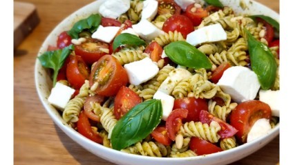 A bowl of colorful pasta salad featuring fusilli pasta, cherry tomatoes, diced mozzarella, fresh basil leaves, and pine nuts, all mixed with a vibrant green pesto sauce. The dish is served on a wooden table.