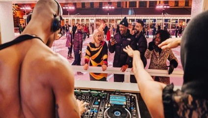 A shirtless DJ stands at a mixing table playing music. In the background, people bundled up in winter clothes, including a woman in a black and yellow sweater, are ice skating and smiling. One woman raises her hands in excitement. The scene is lively and festive.