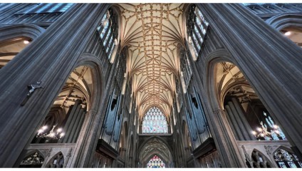 A grand Gothic cathedral interior featuring tall, pointed arches, intricate ribbed vaulting on the ceiling, and large stained glass windows. The columns are adorned with decorative details, and mounted lights illuminate the space.